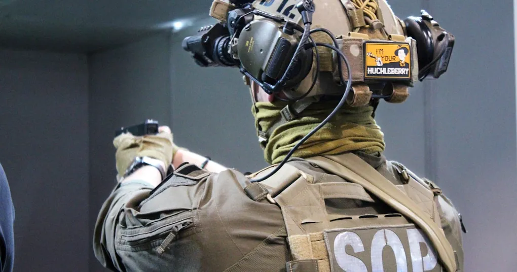 A photo of a US military personnel using a GPS device, with a blurred background of military equipment and vehicles.